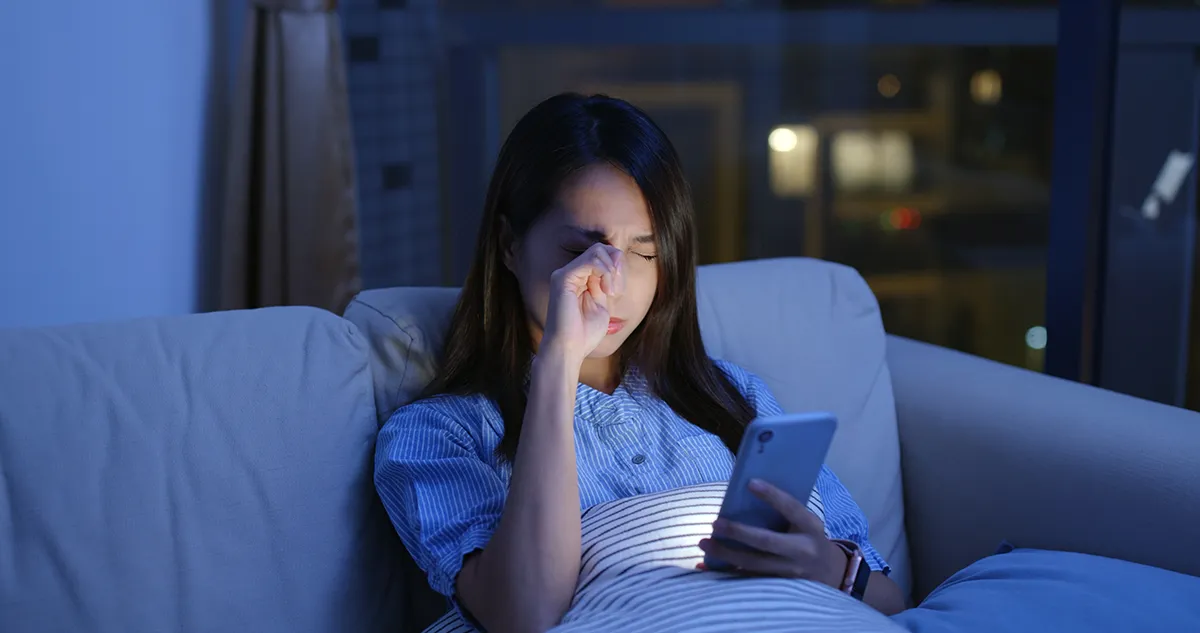 a woman sitting on her sofa at night with her phone and rubbing her eye