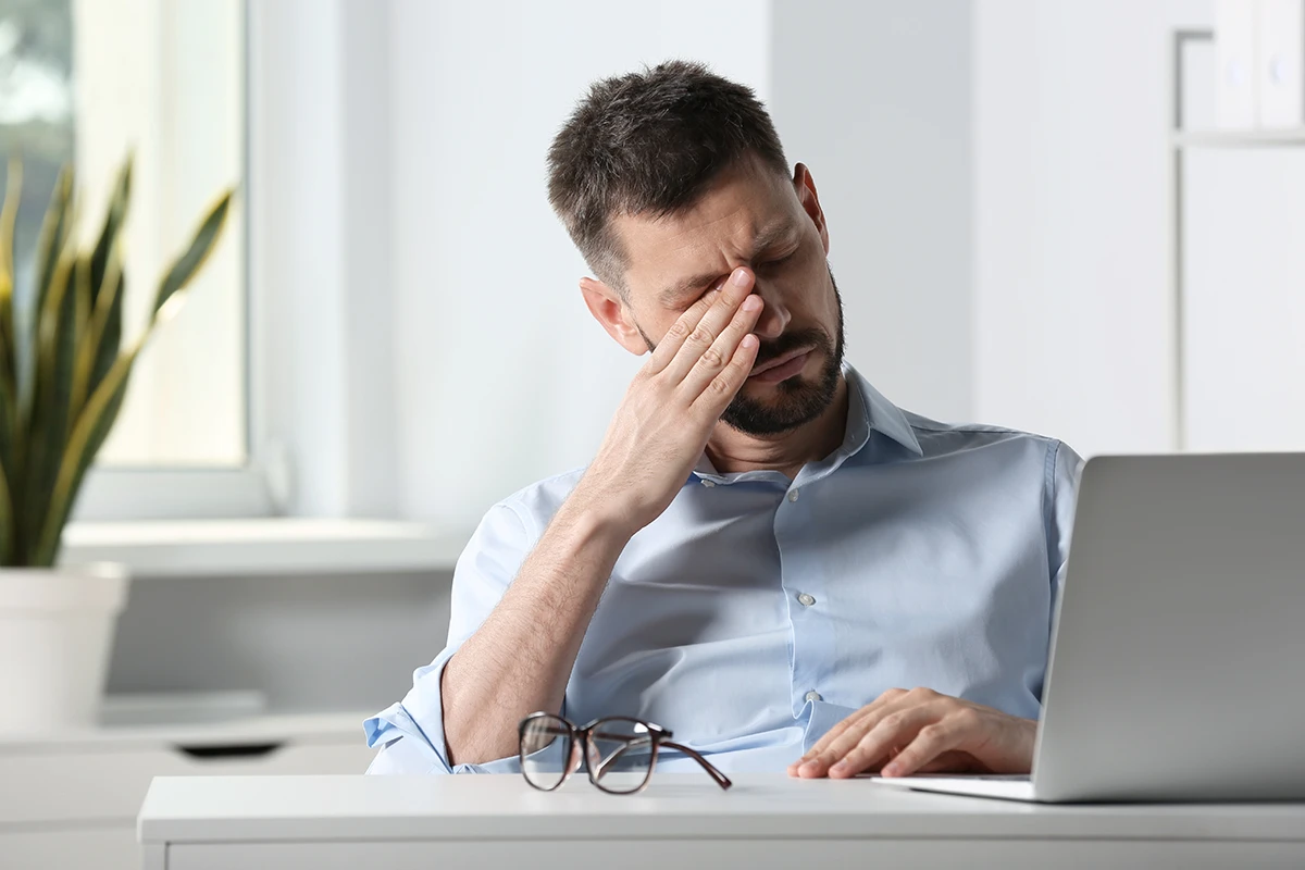 a man sitting at his desk with his glasses off and rubbing his eye