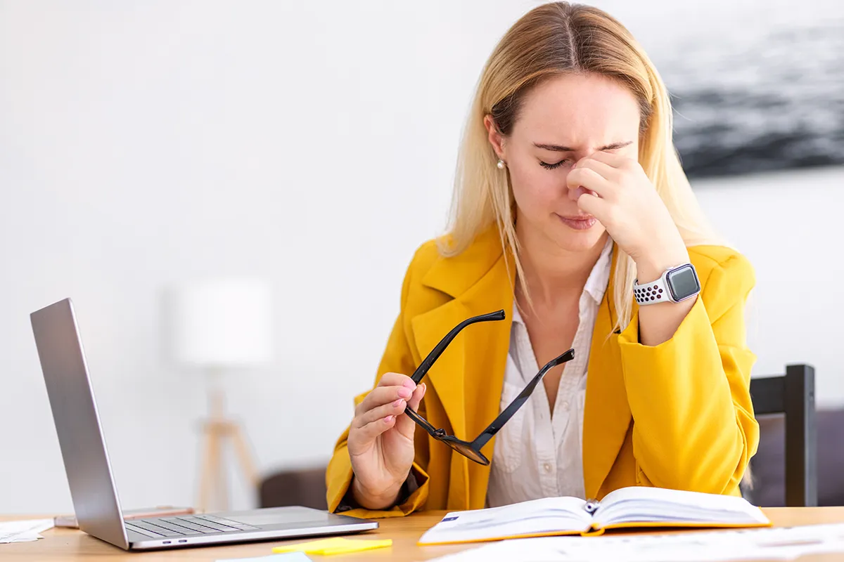 a woman taking off her glasses at her desk and looking uncomfortable
